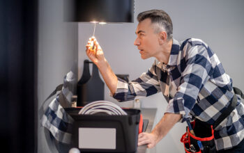 Renovation. Profile of concentrated middle aged man in plaid shirt touching screwdriver to lamp on kitchen range hood in modern kitchen at home